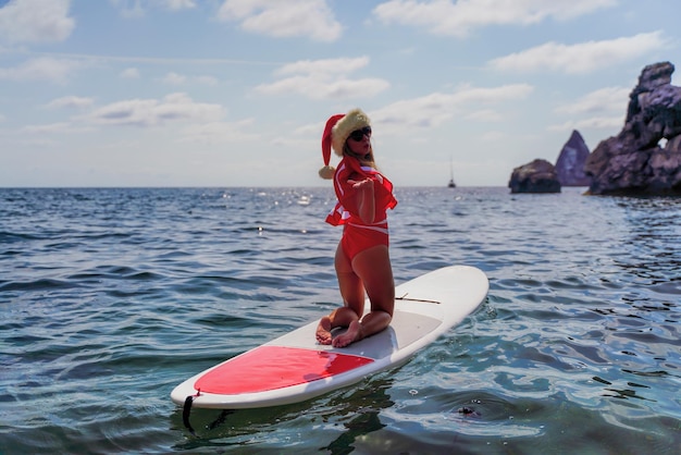 Feliz joven atractiva mujer morena en traje de baño rojo y sombrero de santa nadando a bordo de sup en el mar