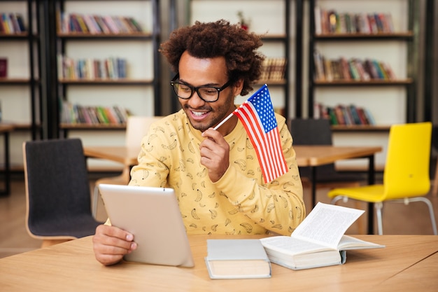Feliz joven con bandera de Estados Unidos sonriendo y usando tableta en biblioteca