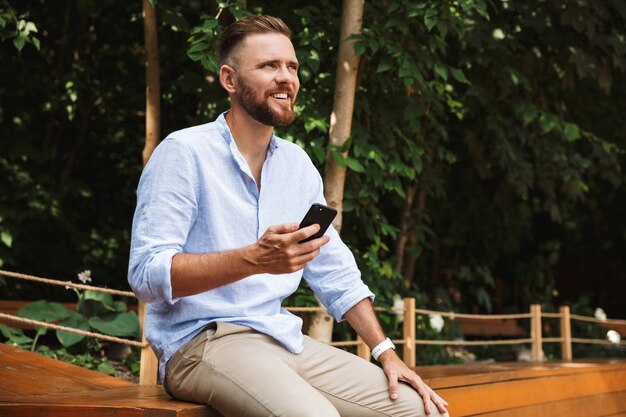 Foto feliz joven barbudo al aire libre mediante teléfono móvil.