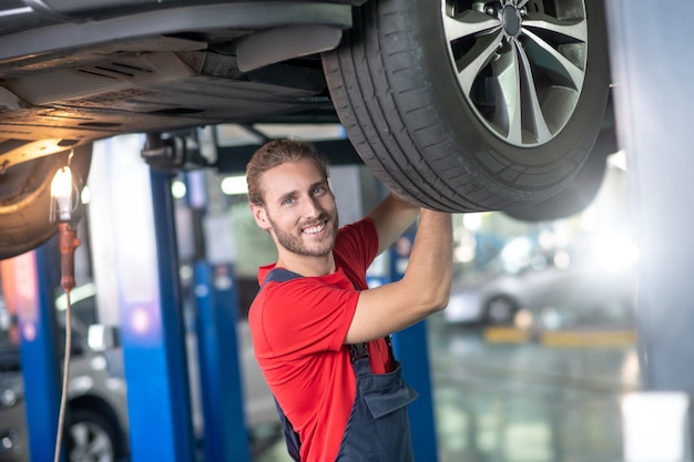 Feliz joven barbudo con un mono de pie bajo el coche levantado trabajando cerca de la rueda