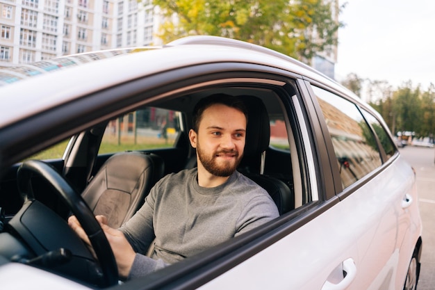 Feliz joven barbudo sentado en el auto y usando la vista lateral del teléfono celular mirando hacia otro lado