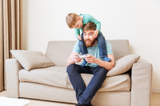 Feliz joven barbudo sentado en el sofá con su hijo sobre los hombros y jugando juegos de computadora