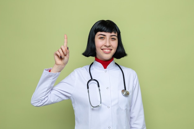 Feliz joven bastante caucásica en uniforme médico con estetoscopio apuntando hacia arriba