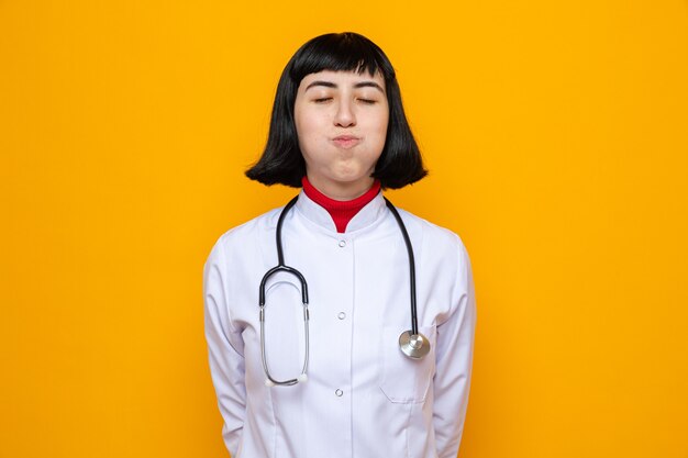 Feliz joven bastante caucásica en uniforme médico con estetoscopio de pie con los ojos cerrados y soplar sus mejillas