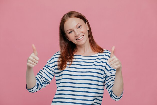 Feliz joven y bella dama europea da doble pulgar hacia arriba, le gusta la idea de alguien o da su aprobación