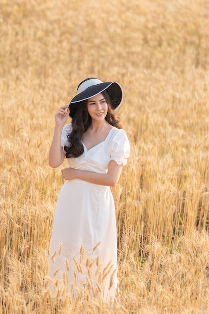 Feliz joven bella mujer asiática en vestido blanco y sombrero de paja de pie en medio del campo de cebada en una tarde soleada
