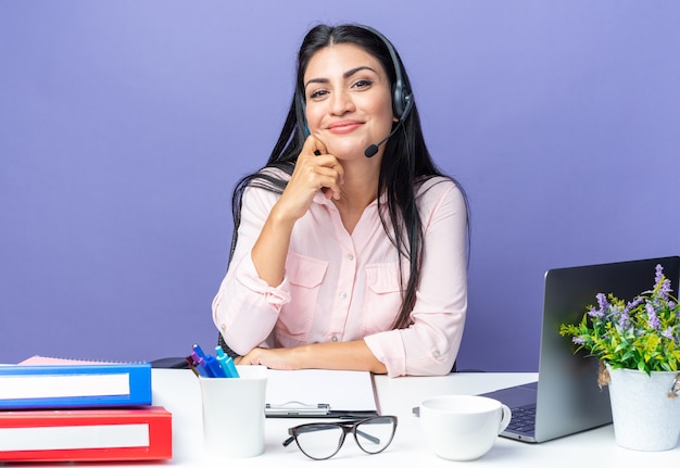 Feliz joven y bella mujer en ropa casual con auriculares con micrófono sonriendo confiado sentado en la mesa con el portátil sobre la pared azul trabajando en la oficina