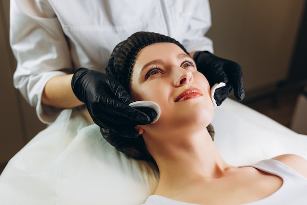 Feliz joven y bella mujer sonriendo con alegría mientras esteticista profesional limpia su rostro