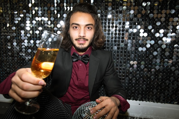 Feliz joven bien vestido animando con flauta de champán mientras está sentado en el suelo junto a la pared brillante en la fiesta