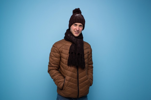 Foto feliz joven con bufanda marrón y gorro de invierno, chaqueta marrón claro sobre fondo azul.