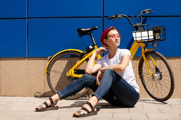 Feliz joven con cabello rosado camina por la ciudad en bicicleta en el verano.