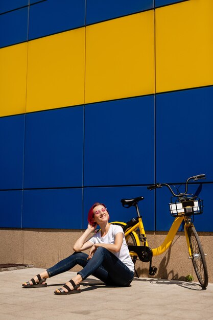 Feliz joven con cabello rosado camina por la ciudad en bicicleta en el verano.