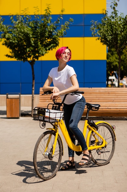 Feliz joven con cabello rosado camina por la ciudad en bicicleta en el verano.