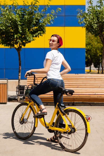 Foto feliz joven con cabello rosado camina por la ciudad en bicicleta en el verano.