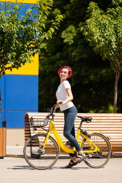 Feliz joven con cabello rosado camina por la ciudad en bicicleta en el verano.