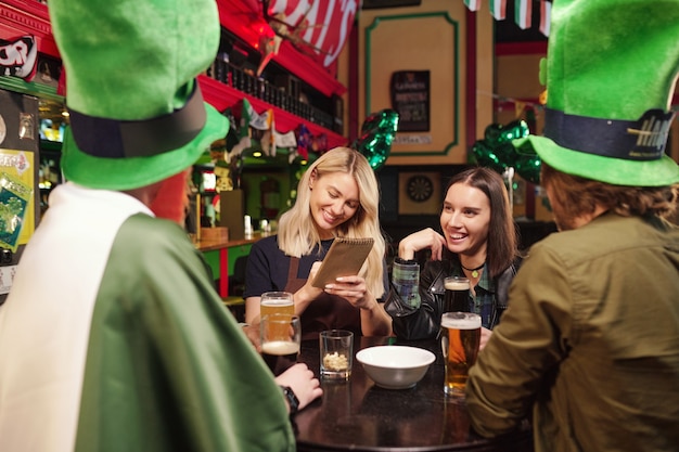 Foto feliz joven camarera rubia en ropa de trabajo escribiendo pedidos de clientes en el bloc de notas entre alegres amigos interculturales con sombreros verdes