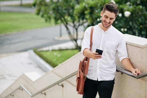 Feliz joven con camisa blanca de pie al aire libre y leyendo un mensaje de texto en el teléfono inteligente