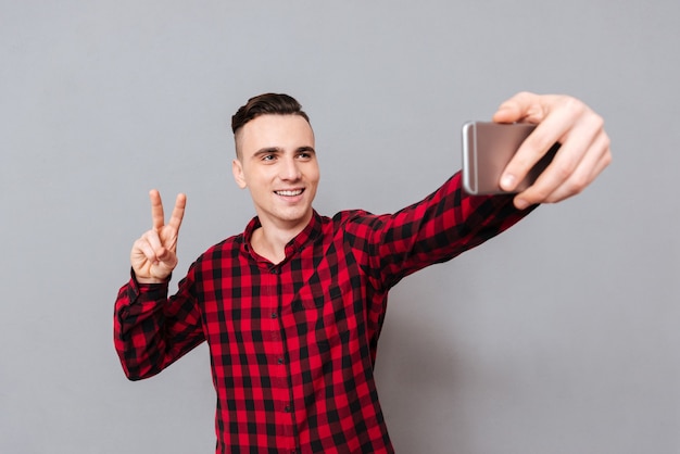 Feliz joven en camisa haciendo selfie y mostrando el signo de la paz. Fondo gris aislado