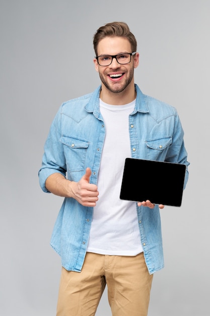 Feliz joven con camisa de jeans de pie y sosteniendo la almohadilla de la pc de la tableta sobre la pared gris del estudio