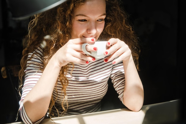 Feliz joven caucásica bebiendo café mientras ve un video en una computadora portátil en casa