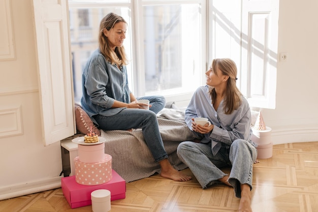 Feliz joven caucásica junto con su hermana charlando y sosteniendo tazas de café en casa. concepto de unidad