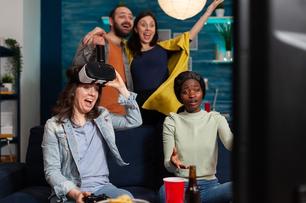 Feliz joven celebrando la victoria mientras juega videojuegos con casco de realidad virtual y sus amigos multiétnicos la animan mientras socializan por la noche.