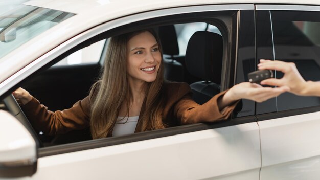 Feliz joven clienta sentada dentro de un auto nuevo tomando la llave automática del vendedor en el moderno