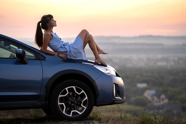 Foto feliz joven conductora con vestido azul disfrutando de una cálida noche de verano recostada sobre el capó de su auto concepto de viaje y vacaciones