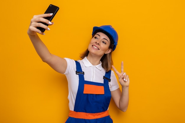Feliz joven constructor mujer en uniforme de construcción y casco de seguridad sonriendo alegremente mostrando v-sign haciendo selfie con smartphone