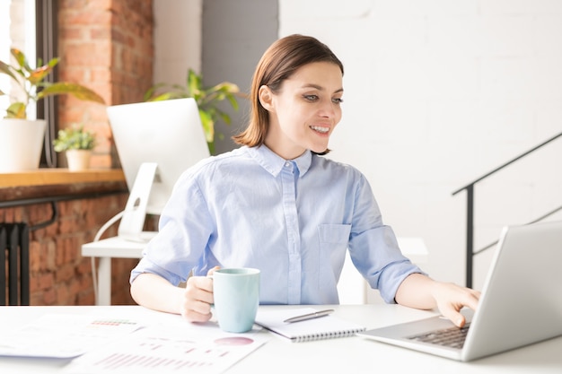 Feliz joven corredor femenino exitoso en camisa mirando la pantalla del portátil mientras navega en la red durante la pausa para el café en la oficina