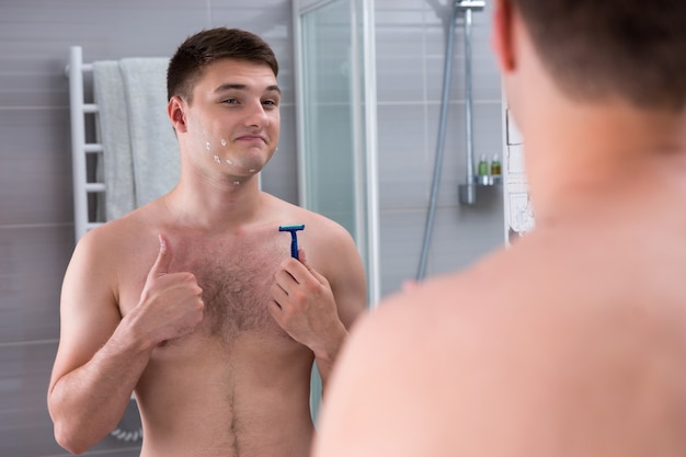 Feliz joven con cortes en la cara sosteniendo una navaja mientras está de pie frente al espejo en el moderno baño de azulejos en casa