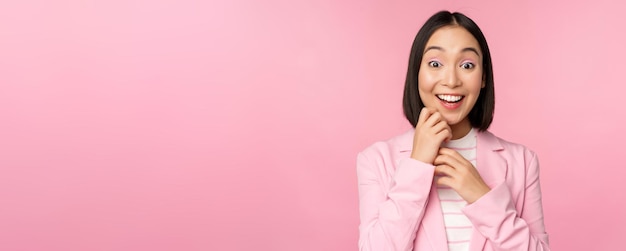 Feliz joven dama de oficina mujer de negocios coreana con traje que parece sorprendida por la cámara posando contra el fondo del estudio en rosa