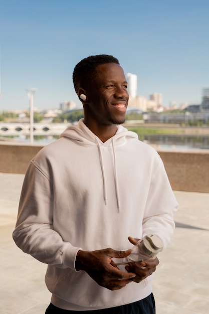 Feliz joven deportista africano con una gran sonrisa sosteniendo una botella de agua mientras descansa después del entrenamiento al aire libre y escucha música