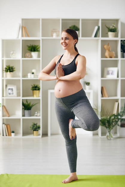 Feliz joven deportista embarazada de pie sobre una pierna en la estera con las manos juntas por el pecho durante el entrenamiento de yoga en casa