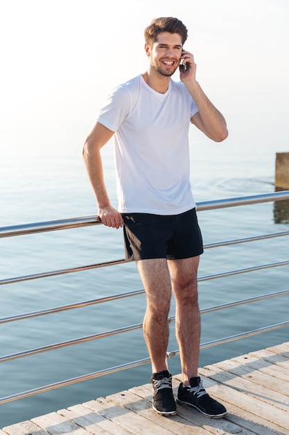 Feliz joven deportista de pie en el muelle y hablando por teléfono móvil