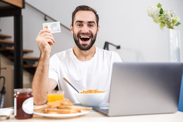 Feliz joven desayunando en la cocina, usando la computadora portátil, mostrando la tarjeta de crédito