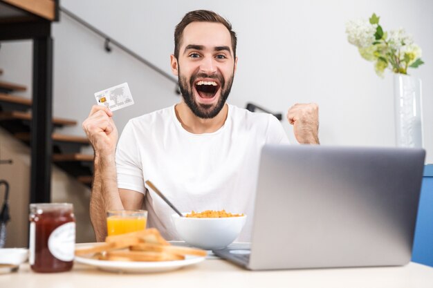 Feliz joven desayunando en la cocina, usando la computadora portátil, mostrando la tarjeta de crédito