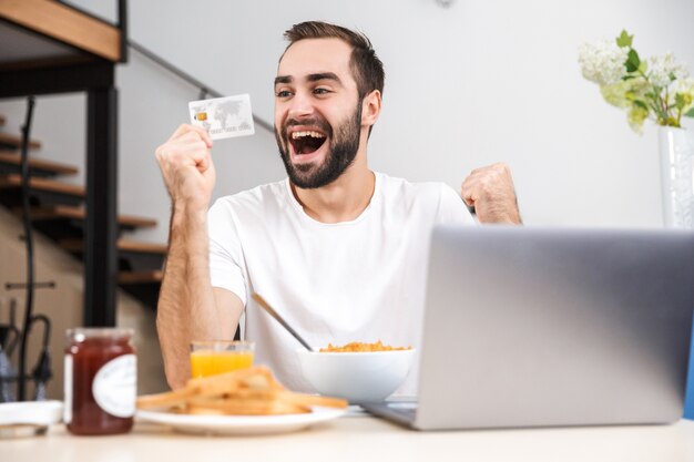 Feliz joven desayunando en la cocina, usando la computadora portátil, mostrando la tarjeta de crédito