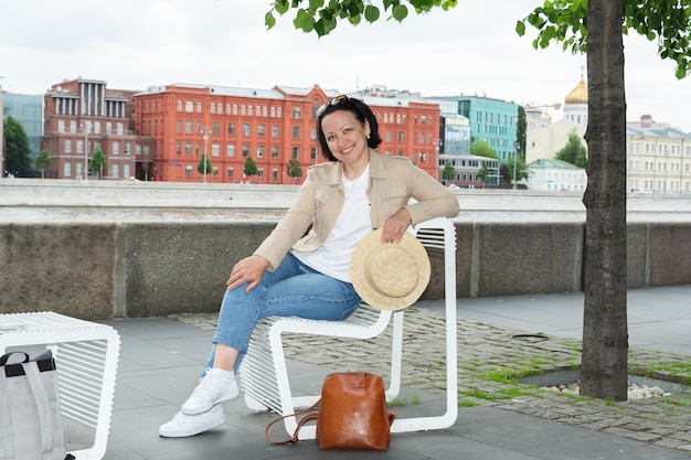 Foto feliz joven está descansando en un banco en el terraplén del río moscú en verano y sonriendo.