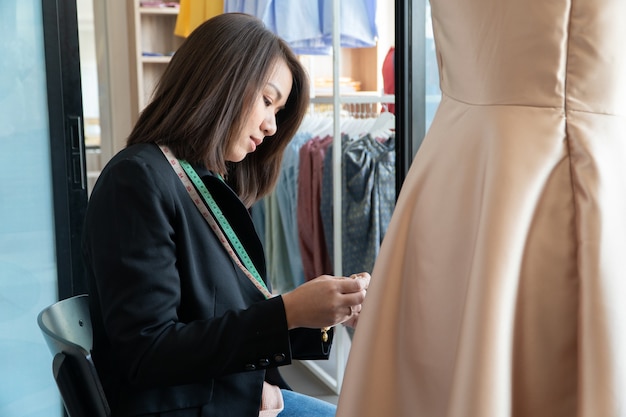 Feliz joven diseñadora de moda modista asiática está comprobando la finalización de un vestido