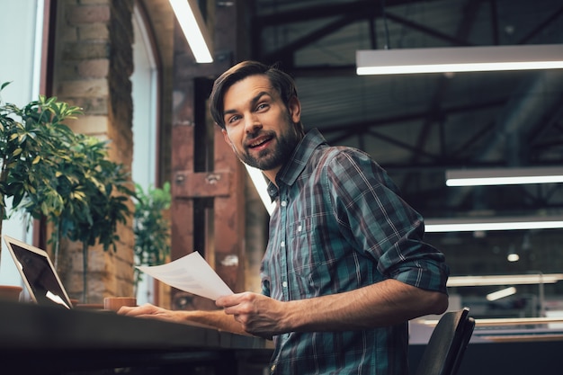 Foto feliz joven con un documento en la mano sentado en la mesa y girando desde su computadora portátil con una sonrisa de satisfacción