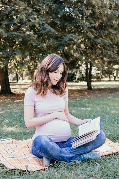 Feliz joven embarazada leyendo un libro en el parque en un día de primavera o verano.