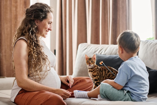 Feliz joven embarazada mirando a su lindo hijo jugando con gato mascota