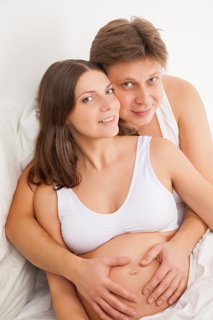Feliz joven embarazada con su marido en la cama blanca en el dormitorio de luz