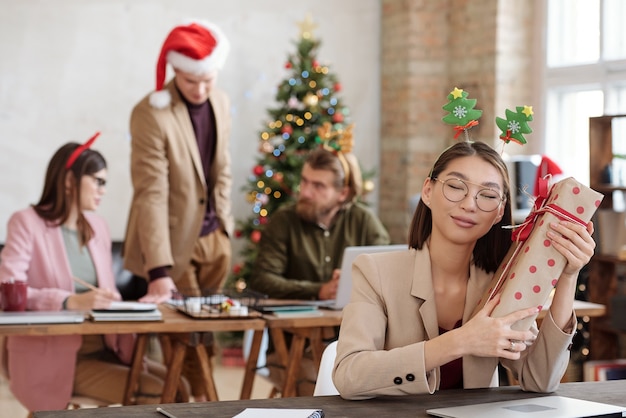 Feliz joven empresaria asiática en diadema navideña sentada junto a la mesa y sosteniendo una gran caja de regalo junto a la oreja mientras mantiene los ojos cerrados