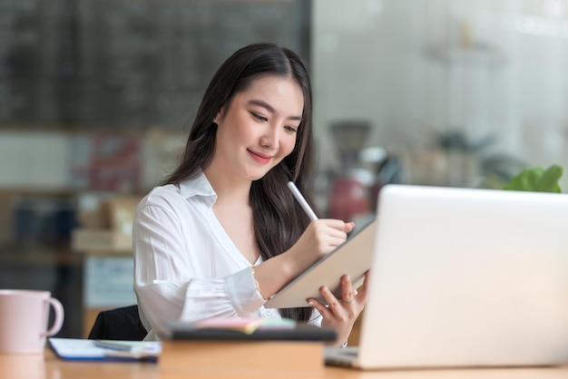 Feliz joven empresaria asiática trabajando en una laptop y tomando notas en la oficina