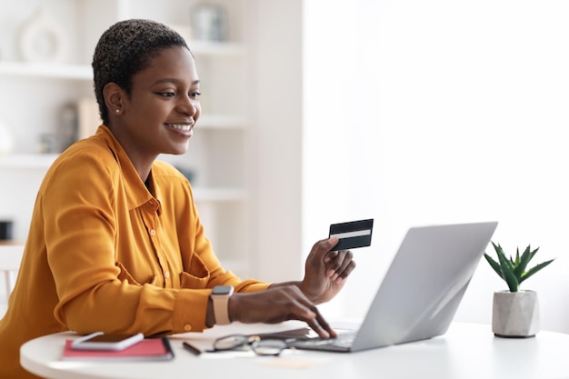 Feliz joven empresaria negra usando laptop con tarjeta de crédito