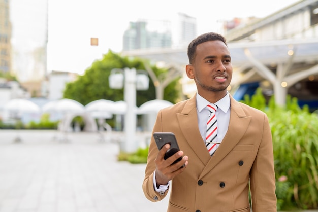 Feliz joven empresario africano guapo pensando mientras usa el teléfono en la ciudad