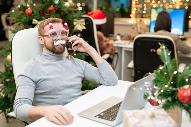 Feliz joven empresario en anteojos de Navidad felicitando a los socios comerciales por teléfono en el lugar de trabajo en la oficina