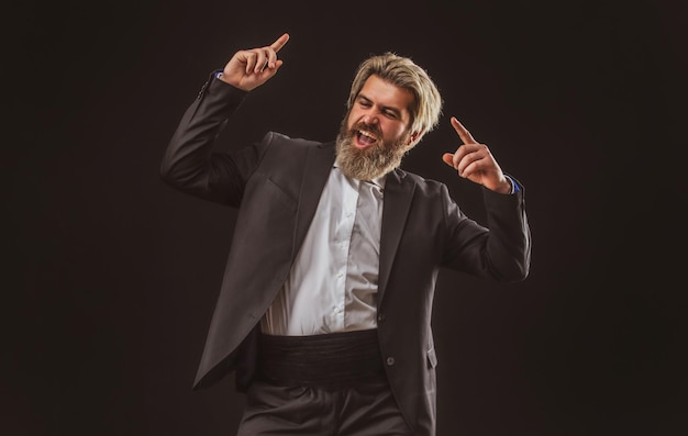 Feliz joven empresario atractivo en traje negro escuchando música con auriculares y bailando sobre gris